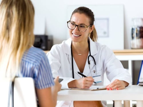 Female doctor smiling during a consultation, holding a clipboard and talking to a patient in a healthcare setting.