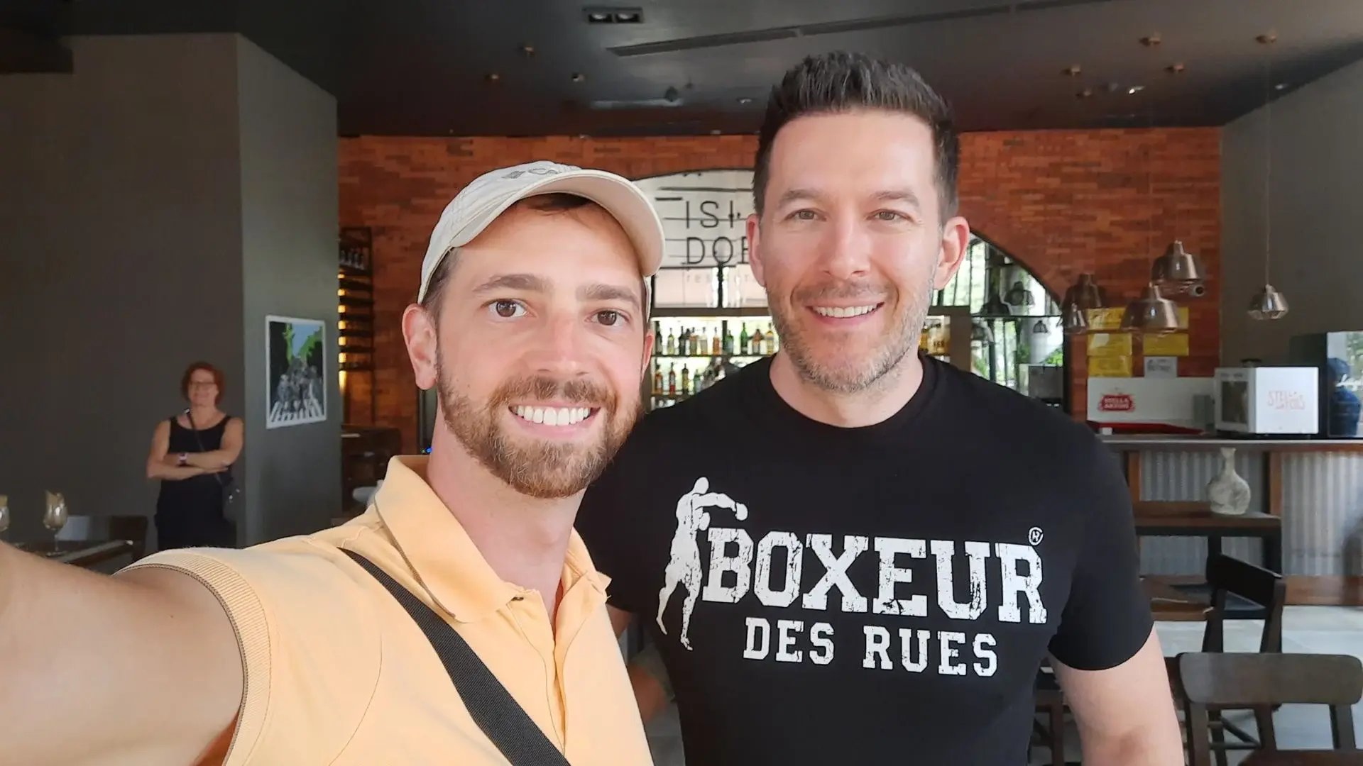 Mikkel Thorup in a beige cap and polo shirt taking a selfie with another man wearing a black “Boxeur des Rues” t-shirt, both smiling in a casual indoor setting