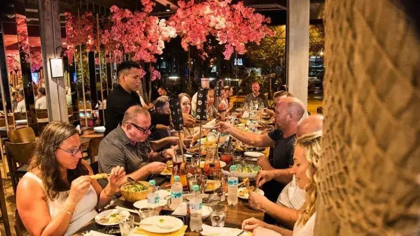 A long table of people enjoying a meal together in a warmly lit restaurant with cherry blossom-style decor overhead.