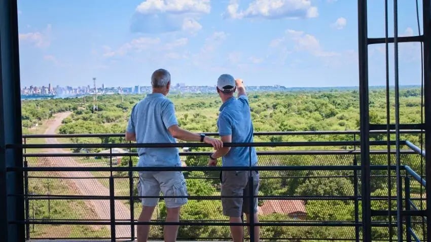 Two men standing on a balcony pointing at a panoramic view of green fields and a distant city skyline under a bright blue sky.