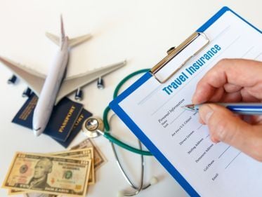 Close-up of a travel insurance form on a clipboard, with passports, cash, a stethoscope, and a toy airplane symbolizing international travel and coverage.