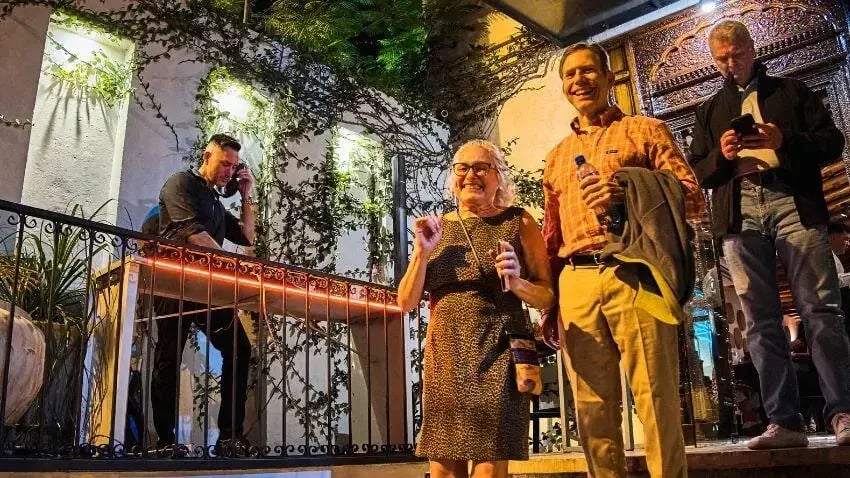 A woman and man smiling outside a venue at night, with others in the background, illuminated by decorative lights and plants.