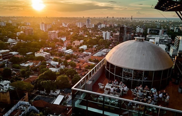 Sunset over Paraguay, viewed from a rooftop, with a cityscape and people gathered on a terrace in the foreground.