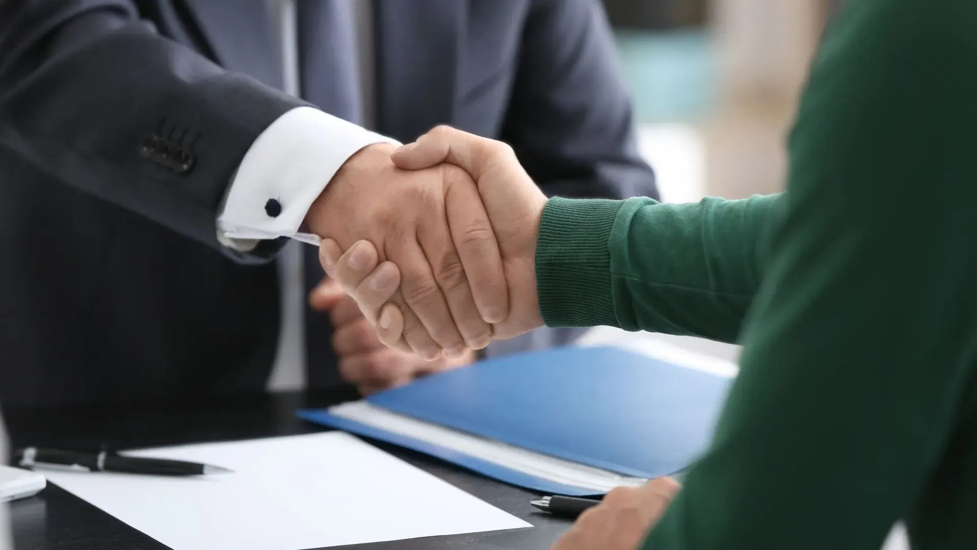 Close-up of a business handshake between two people, symbolizing partnership and agreement, with documents and a pen on the table.
